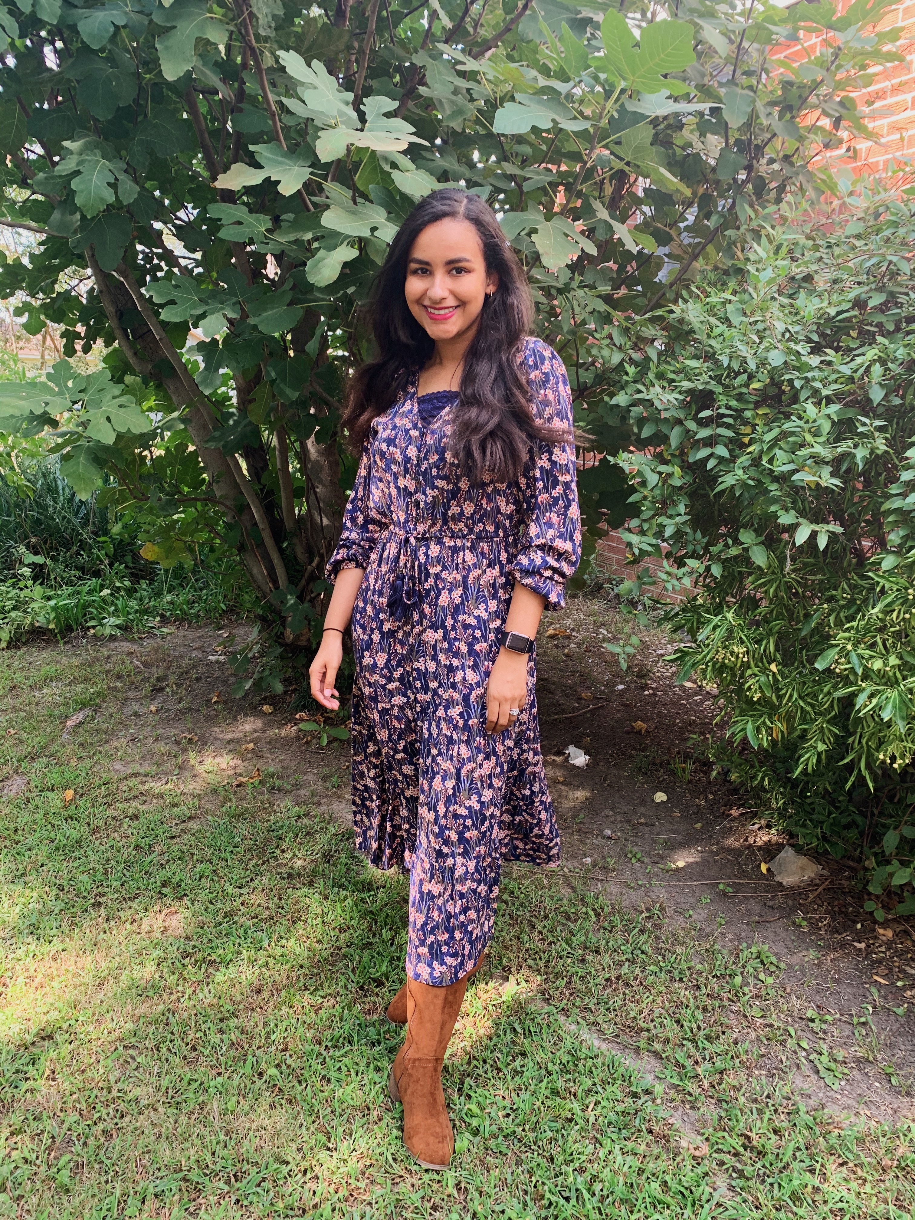 Cassidy standing in front of a tree beside her house. She is wearing a long sleeve dress with brown boots.